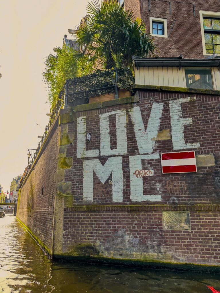 Love Me street art on historic brick wall on the canal in Amsterdam with lush green and red and white sign.